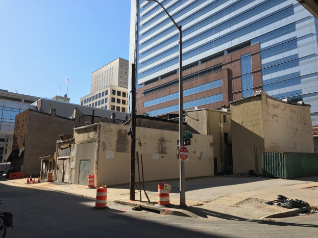 Eine Stadtstraße mit einem geparkten Fahrrad, umgeben von Gebäuden, Straßenmöbeln, Kraftfahrzeugen und einer abgerissenen Gebäude im Hintergrund unter einem klaren blauen Himmel.
