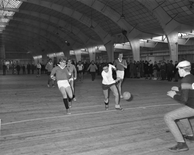 Eine Gruppe von Menschen, die drinnen Fußball auf einem schwarz-weißen Platz spielen, mit einer Wand im Hintergrund und einer Decke mit Lampen oben.