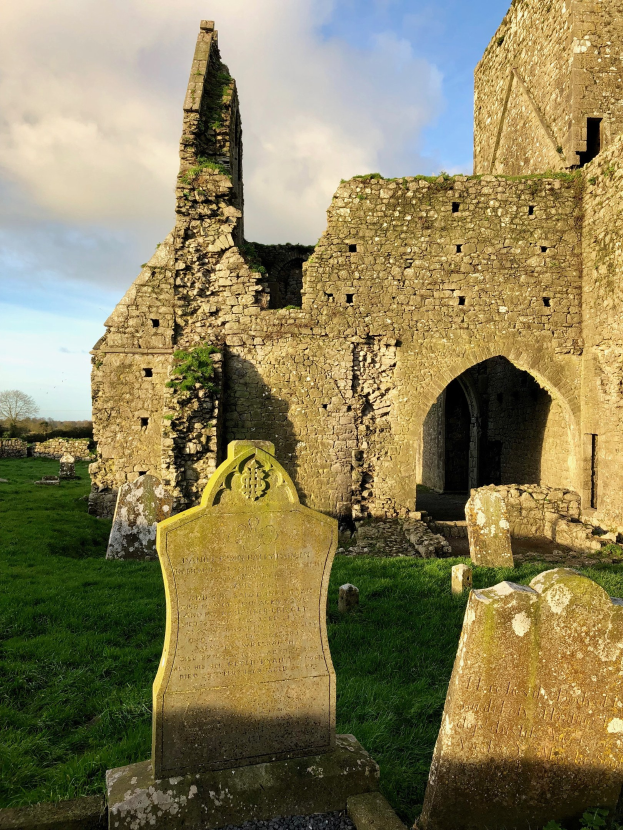 Ruinen einer alten Kirche in Irland mit einem Friedhof im Vordergrund und einer Burg im Hintergrund bei einem bewölkten Himmel, mit sichtbarem Wasserzeichen.
