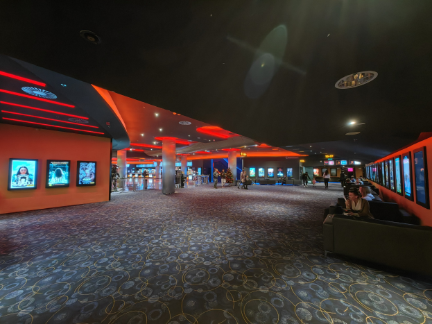 Ein geräumiger Raum mit Menschen auf Sofas sitzend, die Fernsehgeräte an den Wänden anschauen, mit Säulen darin verstreut und Deckenlampen, die den Boden unten zeigen.