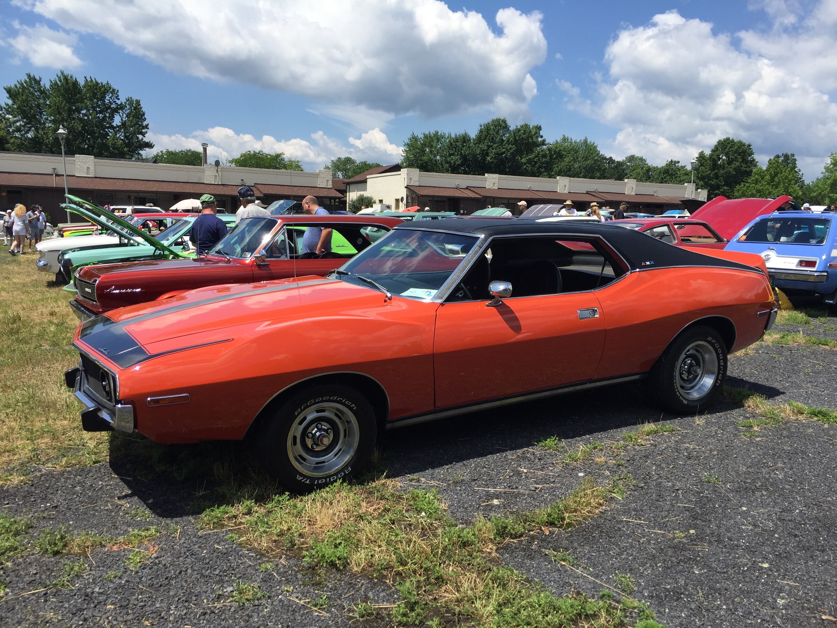 Oldtimer auf einem Grasparkplatz mit Menschen in Kappen und Gebäuden im Hintergrund.