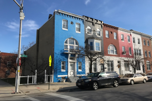 Eine Reihe bunter Häuser an einer Straßenecke in Baltimore, Maryland, mit geparkten Autos, Bäumen, Pfählen, Brettern und einem Zaun, der die Straße säumt, unter einem bewölkten Himmel.