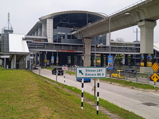 Autobahn mit fahrenden Autos, eine entfernte Brücke, beleuchtete Straßenlaternen, Schilder an Pfählen, grasbewachsener Straßenrand, ein Gebäude mit Säulen, Bäume und ein bewölkter Himmel.