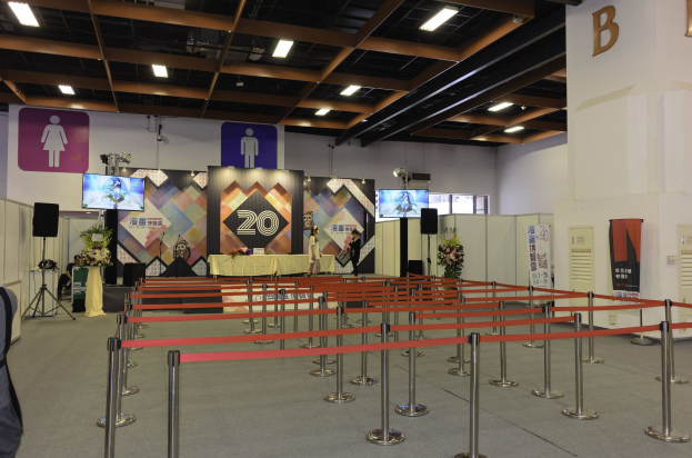 Großer Ausstellungssaal mit Tischen und Stühlen in Reihen, Absperrpoller mit Bändern, Blumensträuße, Rednerpulte, Banner, eine mit dem Buchstaben B markierte Wand und Deckenbeleuchtung.