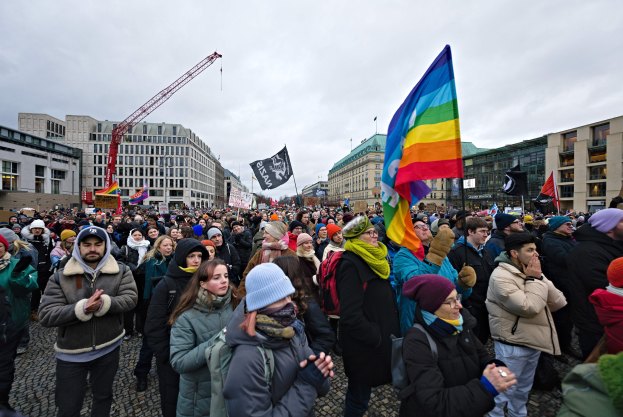 Eine große Gruppe von Menschen mit Fahnen und Transparenten mit der Aufschrift 'Lgbtq+ Rechte Demonstrationen in Berlin', die vor einem Gebäude mit anderen Strukturen und einem bewölkten Himmel stehen.