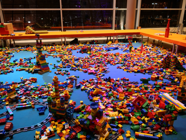 Ein großer Raum mit farbenfrohen Lego-Blöcken, die auf dem Boden verstreut sind, umgeben von Tischen und Säulen, mit Glasfenstern im Hintergrund, die Menschen und Lichter zeigen.