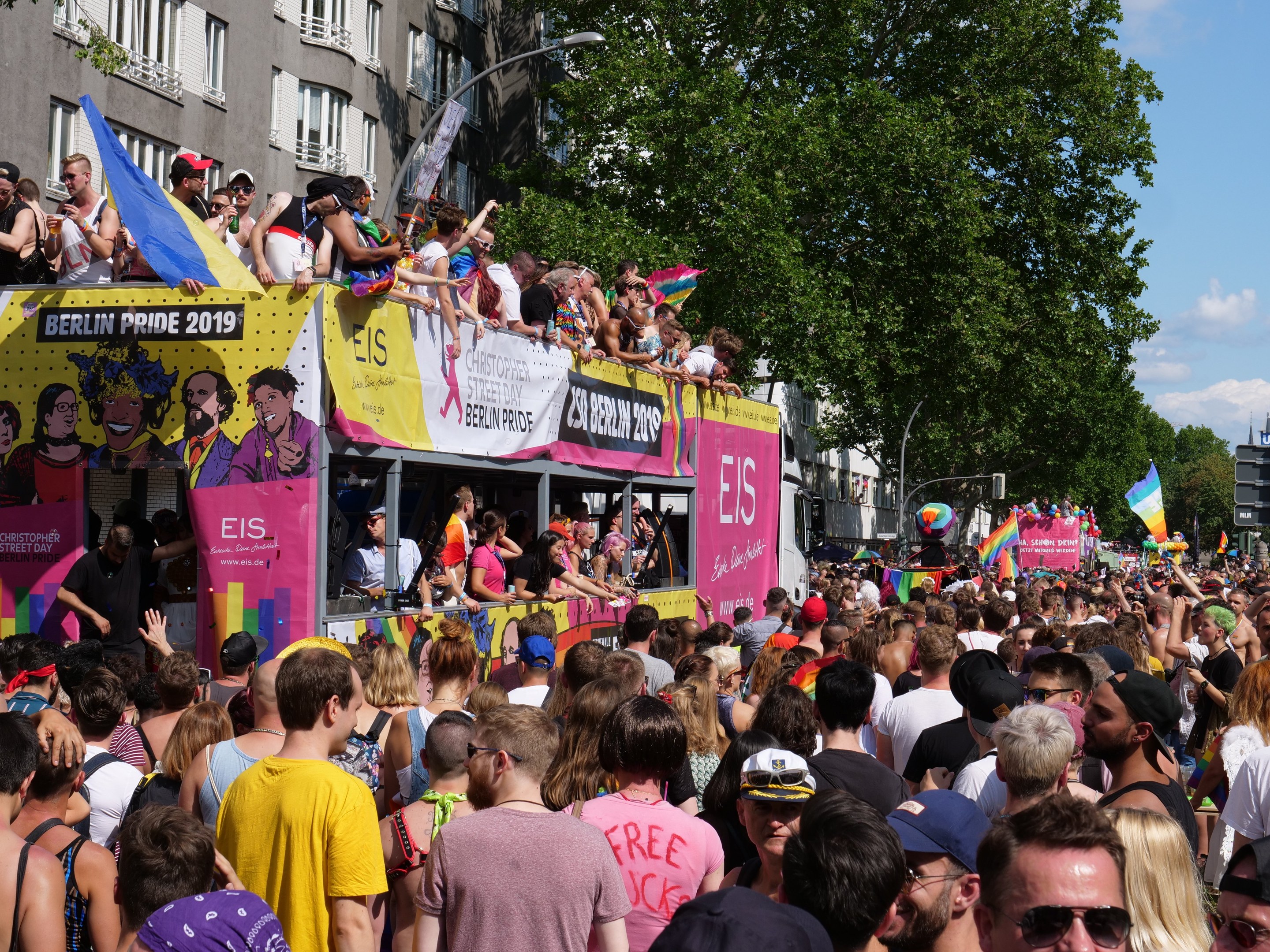 Eine große Menge marschiert während des Berlin Pride 2019 die Straße entlang, viele tragen Kappen und Schutzbrillen, einige halten Fahnen, mit Bannern, Laternenpfählen, Bäumen und Gebäuden im Hintergrund unter einem bewölkten Himmel.