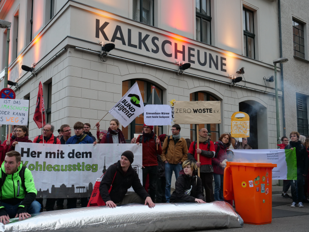 Eine Gruppe von Menschen mit Schildern und Plakaten steht vor einem Gebäude, mit zwei Personen im Vordergrund und einem Müllcontainer rechts, während einer Demonstration in Deutschland, mit Gebäuden, Fenstern, Lichtern und Schildern im Hintergrund.