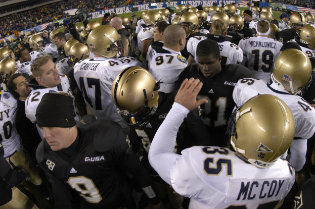 Eine Gruppe von Fußballspielern in Helmen, die sich auf einem Rasenfeld mit Bannern im Hintergrund versammeln, umgeben von Zuschauern in den Rängen.