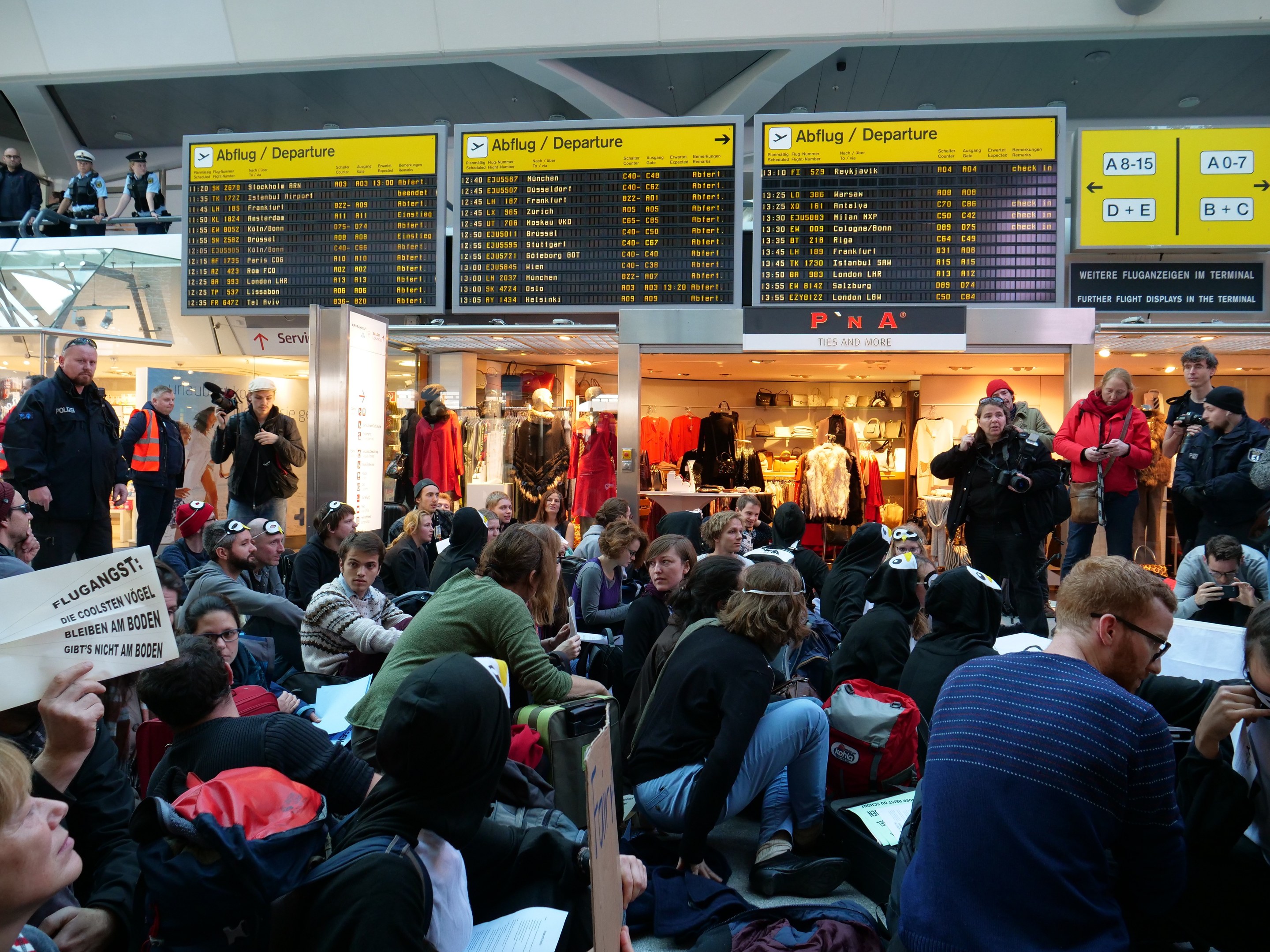 Menschen sitzen und stehen in einem Flughafen während einer Protestaktion, mit Informationsschildern, Schaufensterpuppen in Kleidern und Deckenbeleuchtung im Hintergrund.