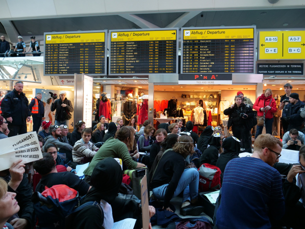 Menschen sitzen und stehen in einem Flughafen während einer Protestaktion, mit Informationsschildern, Schaufensterpuppen in Kleidern und Deckenbeleuchtung im Hintergrund.