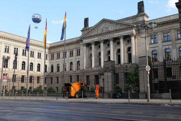 Großes modernes Gebäude mit vielen Fenstern, als Bundestag in Berlin, Deutschland identifiziert, umgeben von Straßeninfrastruktur, Fußgängern und Flaggen, mit Bäumen und einem bewölkten Himmel im Hintergrund.