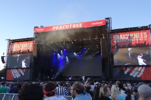 Eine große Menschenmenge steht vor einer hell erleuchteten Bühne beim Shaky Knees Music Festival, auf der Musiker unter Bannern spielen, mit einem Geländer im Vordergrund und einem klaren blauen Himmel dahinter.