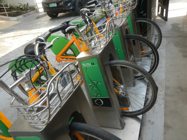 Eine Reihe von Fahrrädern, die in einer Parkanlage mit einem Auto auf der Straße dahinter, einem Tor auf der rechten Seite und Pflanzen und einer Wand auf der linken Seite geparkt sind, Teil eines Shuttle-Fahrradverleihsystems.