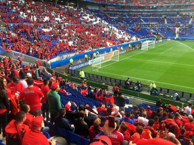 Eine große Menschenmenge in einem Stadion schaut einem Fußballspiel zu, mit Menschen, die auf Stühlen sitzen und stehen, Schildern, einem Torpfosten und Deckenbeleuchtung.