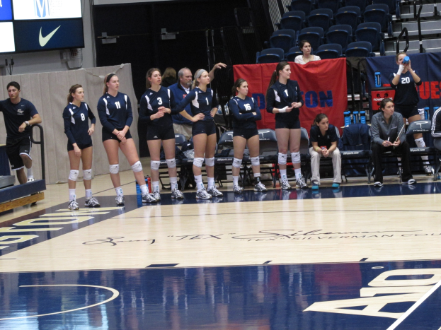 Eine Gruppe von Frauen-Volleyballspielerinnen auf einem Feld, einige sitzen auf Stühlen, mit Bannern, einem Metallzaun, einem Anzeigebildschirm und einer Wand im Hintergrund.