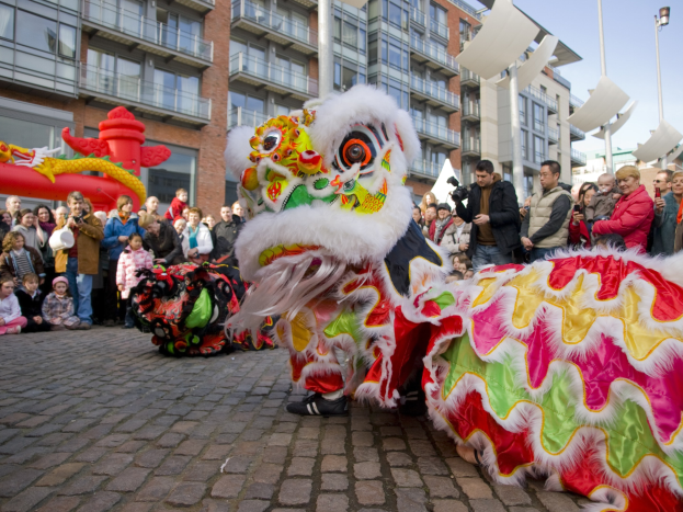 Lebendige Feier des chinesischen Neujahrs in Amsterdam mit einem Löwen tanzen im Vordergrund und einer Menschenmenge, einige mit Kameras, die darum herum versammelt sind, vor einem Hintergrund aus Gebäuden, Laternenmasten und einem klaren blauen Himmel.