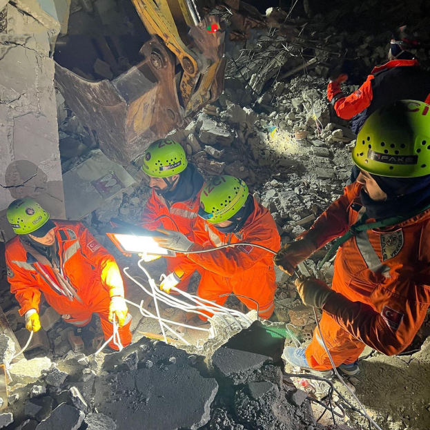 Gruppe von Menschen in orangefarbener Uniform und grünem Helm bei der Arbeit an Schutt mit einem Kran im Hintergrund während einer Rettungsaktion.