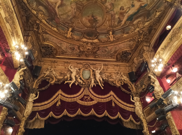 Theaterinnenraum mit roter Gardine, Kronleuchtern, Deckenmalereien, Lichtern und einem Gemälde, das eine Bühne und einige Personen am Boden zeigt.
