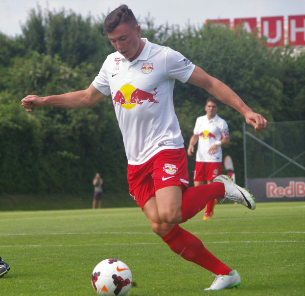 Ein Profifußballer in rot-weißer Uniform kickt einen Fußball auf einem grünen Feld mit Bäumen und einem klaren blauen Himmel im Hintergrund, wobei ein Schild mit der Aufschrift 'RB Leipzig' sichtbar ist.