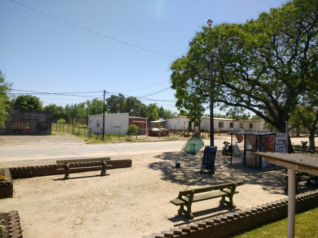 Ein Park mit Bänken, ein Picknicktisch, Gras, blühende Pflanzen, eine Informationstafel, ein Laternenmast, ein Schuppen, eine Straße mit Fahrzeugen, Häuser, ein Zaun, Bäume, Strommasten mit Drähten und eine bewölkte Himmel.