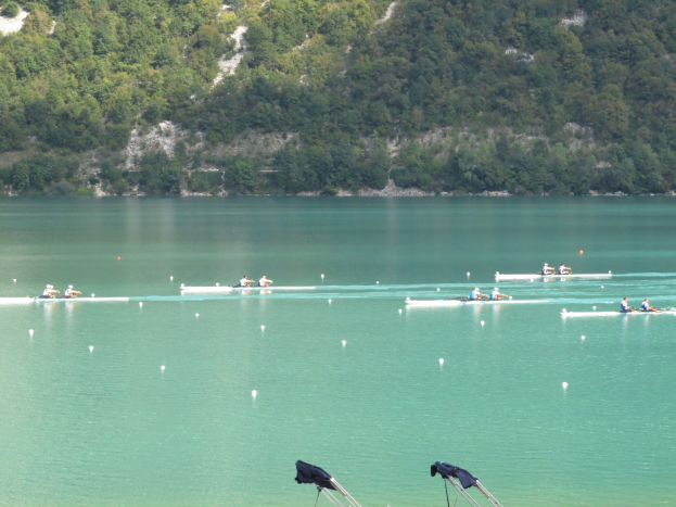 Eine Gruppe von Menschen, die auf einem Gewässer umgeben von Bäumen und einem Hügel rudern, mit Kleiderständern am unteren Bildrand.