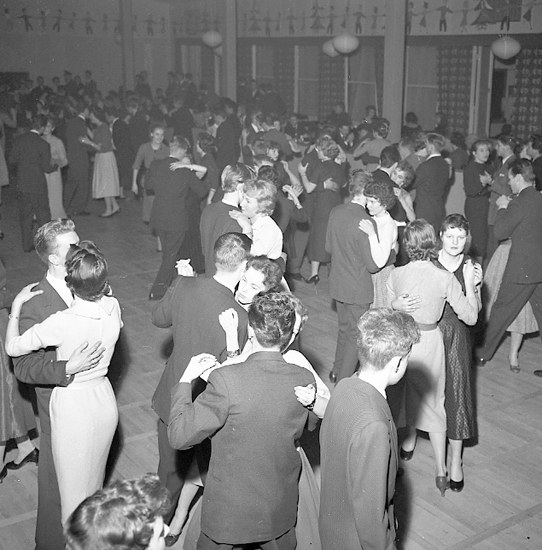 Schwarzes und weißes Foto einer Gruppe von Menschen in Abendgarderobe, die in einem Ballsaal tanzen, mit einer Wand im Hintergrund.