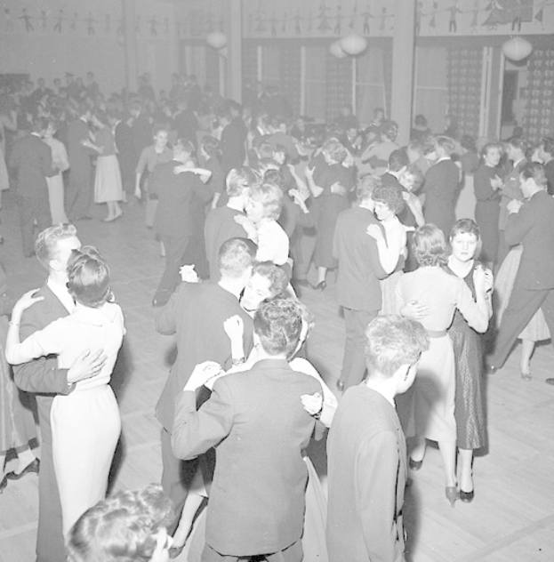 Schwarzes und weißes Foto einer Gruppe von Menschen in Abendgarderobe, die in einem Ballsaal tanzen, mit einer Wand im Hintergrund.