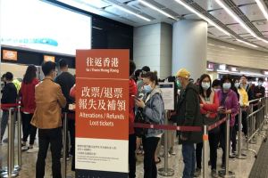 Eine Gruppe von Menschen mit Masken steht in einer Schlange am Hong Kong Airport, umgeben von Absperrpollern mit Bändern, mit einer Tafel im Vordergrund, die "Hong Kong Airport Refunds Due to Covid-19" zeigt, und Säulen und Deckenlampen im Hintergrund.
