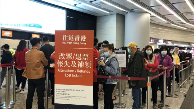 Eine Gruppe von Menschen mit Masken steht in einer Schlange am Hong Kong Airport, umgeben von Absperrpollern mit Bändern, mit einer Tafel im Vordergrund, die "Hong Kong Airport Refunds Due to Covid-19" zeigt, und Säulen und Deckenlampen im Hintergrund.